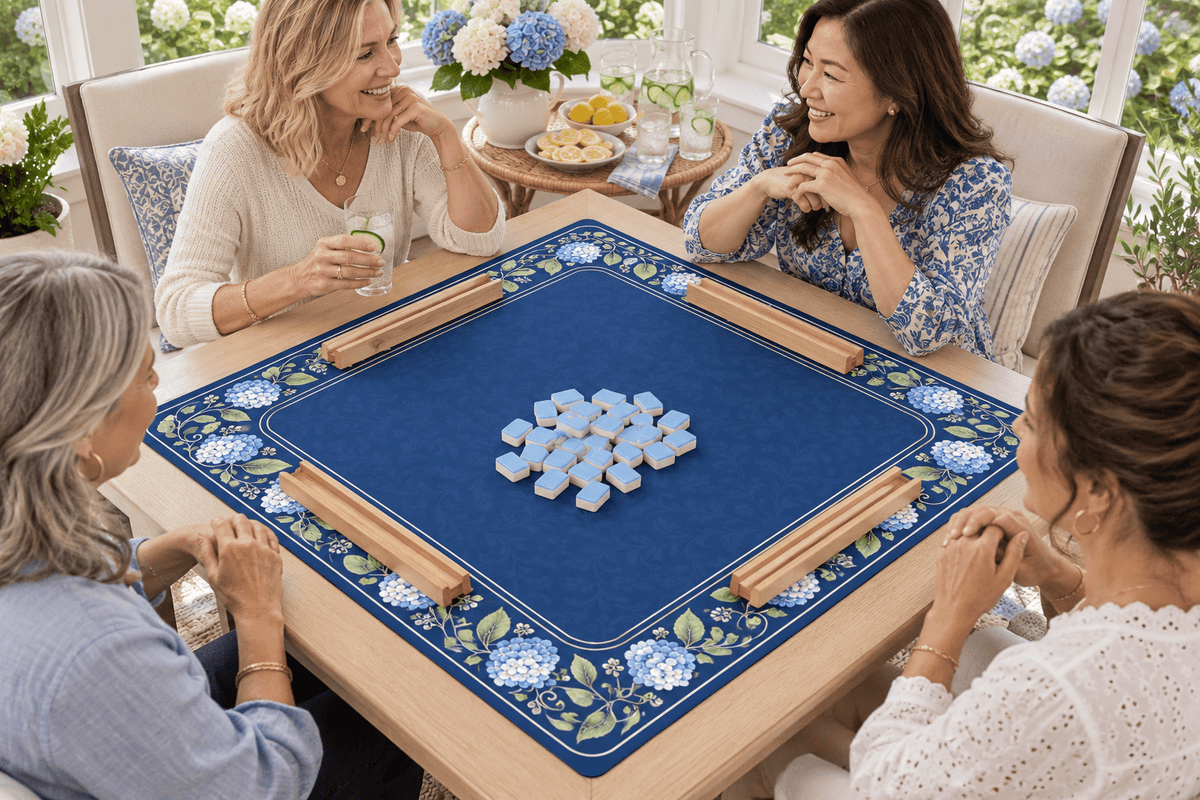 Four friends gathering around a blue HYDRANGEA HOUSE mahjong mat in a bright garden room, with empty wooden racks and face-down tiles before play.
